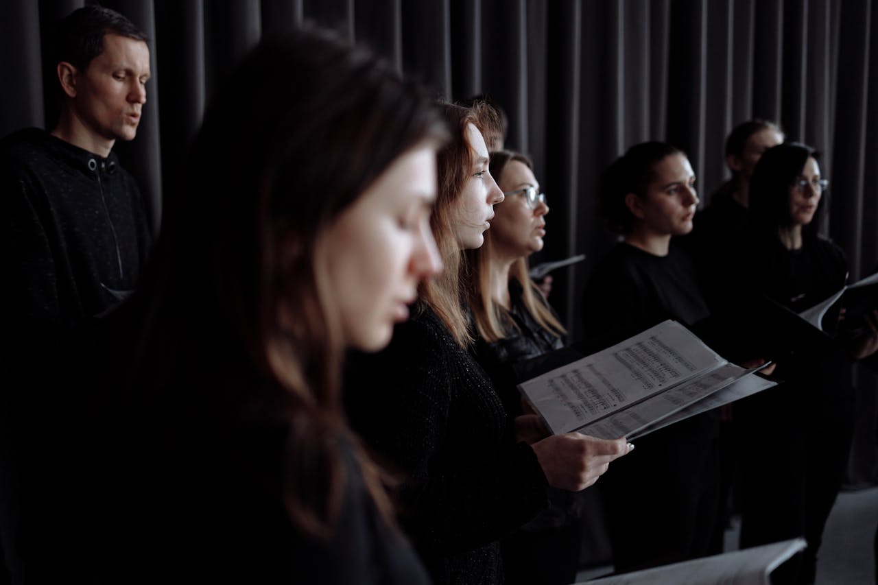 Adult choir practicing in an indoor studio performance setting with music sheets.